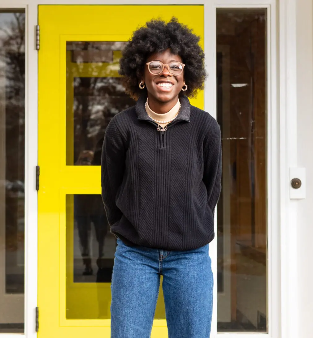 A smiling person with glasses and an afro hairstyle stands in front of a bright yellow door, wearing a black sweater and blue jeans, with their hands behind their back.