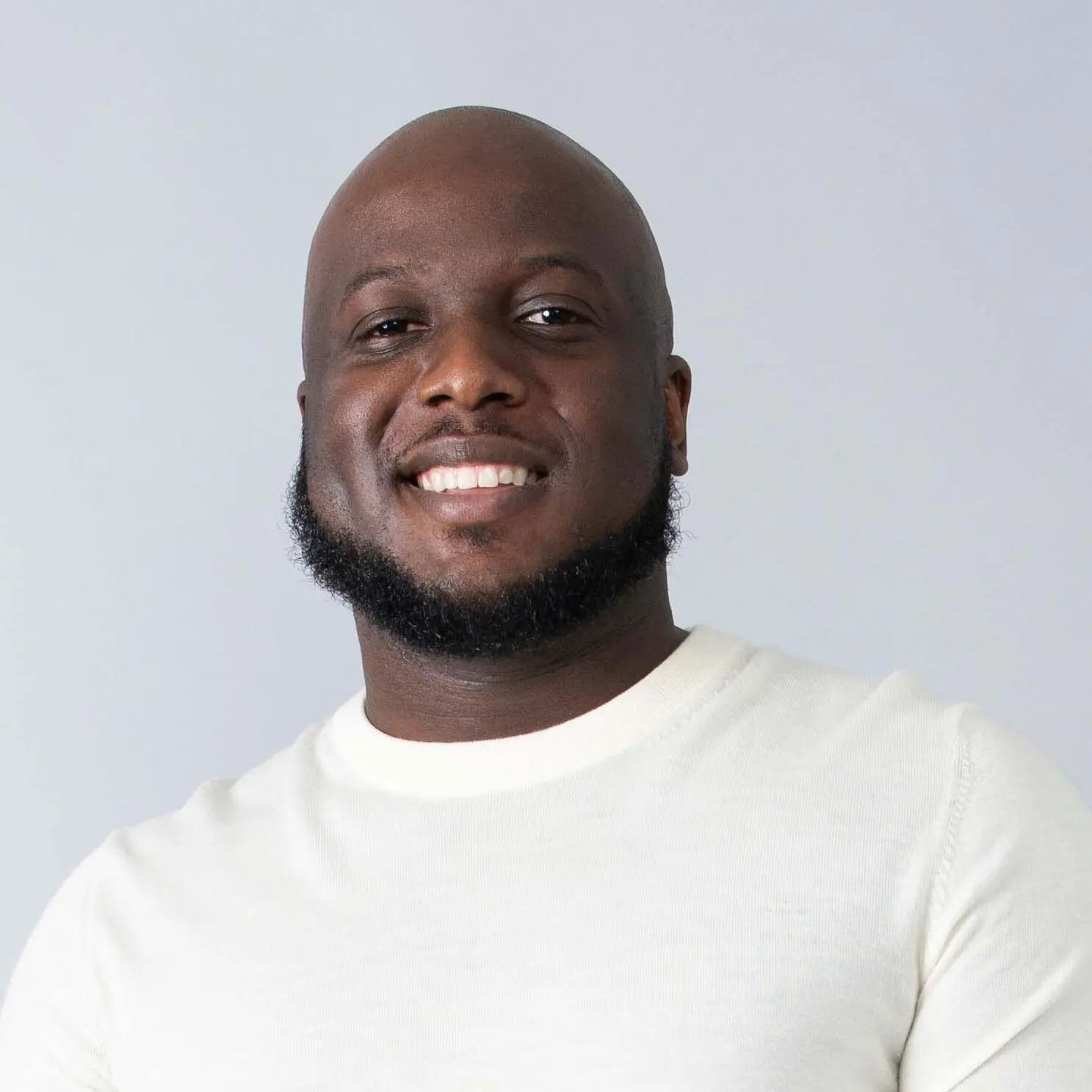 A man with a shaved head and short beard is smiling, wearing a white t-shirt against a light gray background.