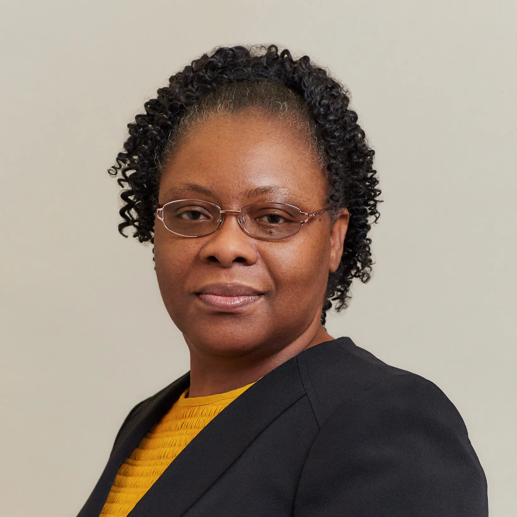 A woman with curly hair, glasses, and a black blazer over a yellow top poses against a plain light background, looking confidently at the camera.