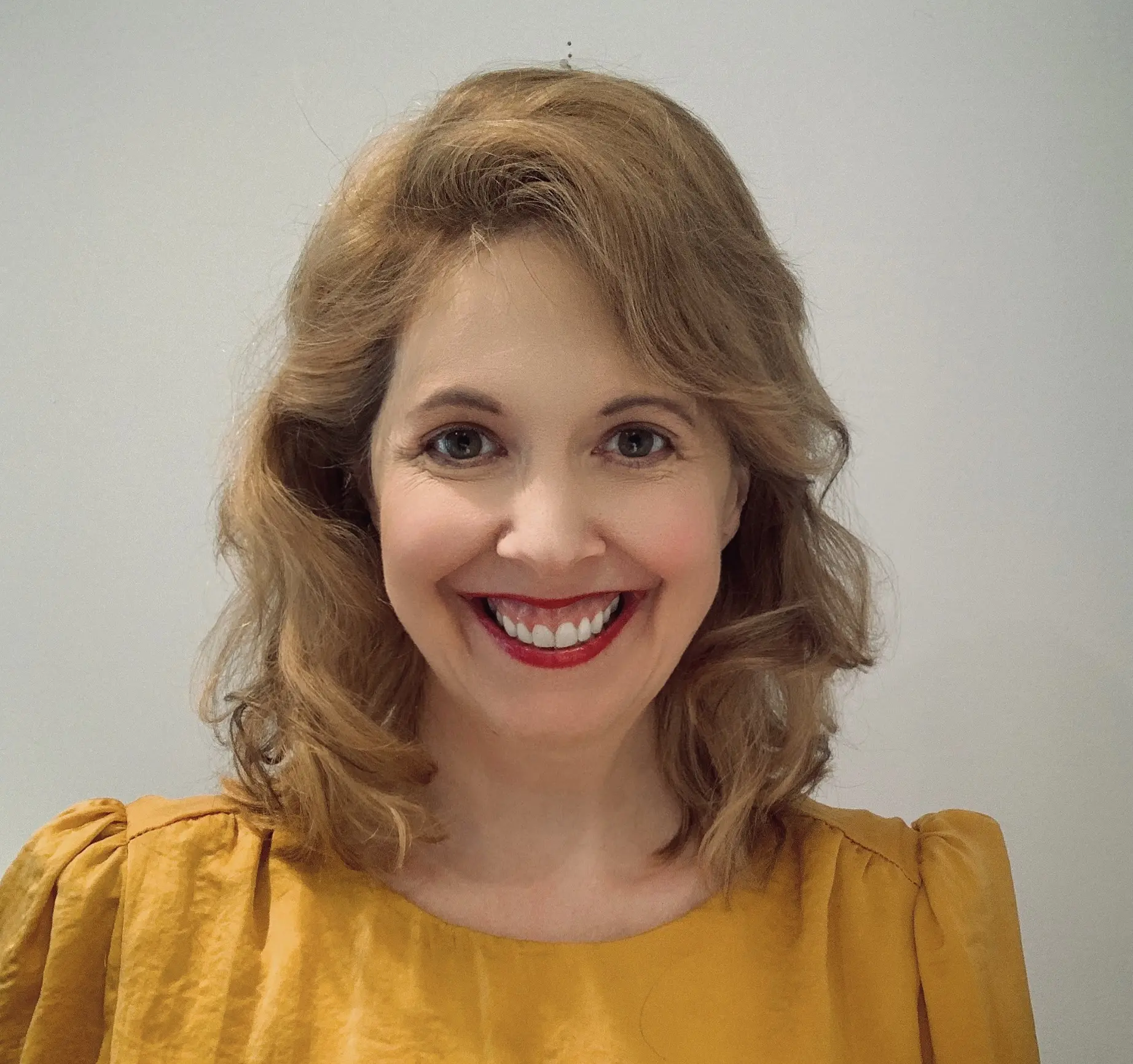 A woman with wavy, shoulder-length blonde hair smiles at the camera. She is wearing a mustard yellow top and has bright red lipstick. The background is plain and light-colored.
