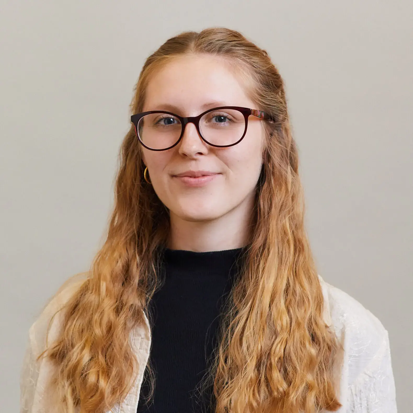 A young woman with long, wavy, light brown hair wears glasses and a black top with a light-colored cardigan. She is smiling softly and posing against a plain, light-colored background.