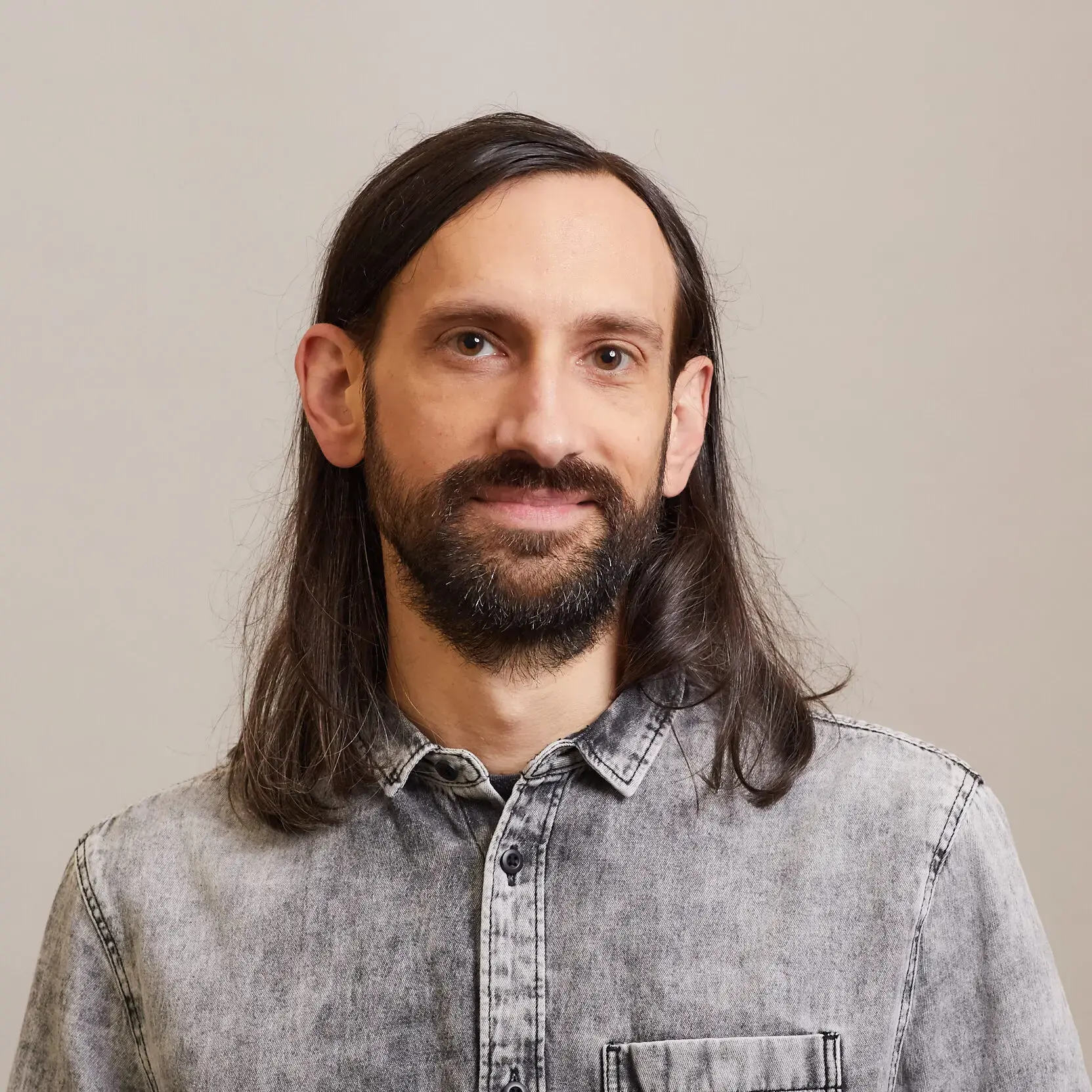 A man with long dark hair and a beard is wearing a light gray button-up shirt. He is standing in front of a plain, light-colored background and smiling slightly at the camera.