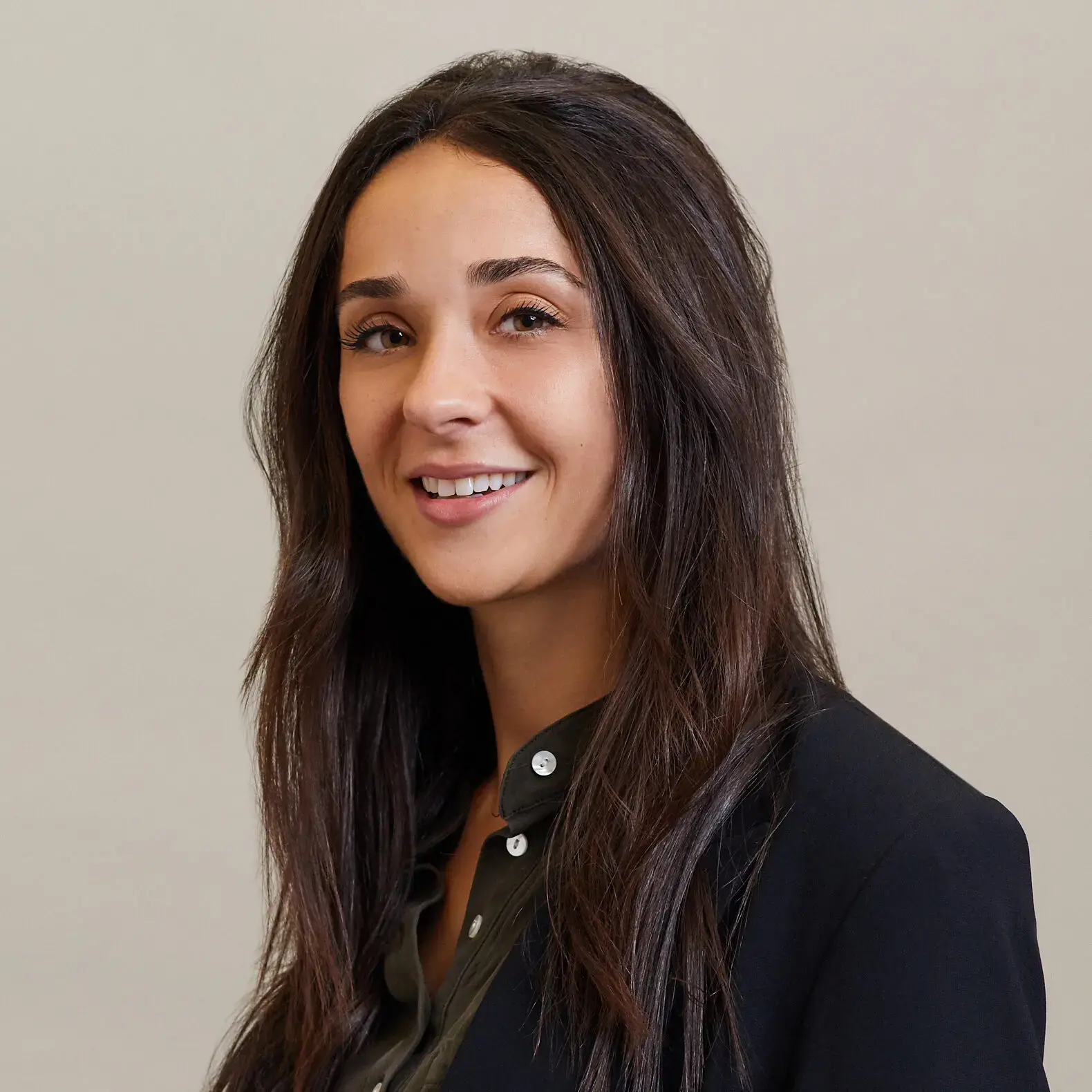 A woman with long brown hair, wearing a dark blazer over a dark shirt, smiles at the camera against a plain light background.