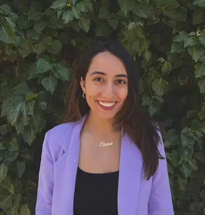 A woman with long brown hair, wearing a lilac blazer, black top, and a necklace that says Ciara, smiles in front of a leafy green background.
