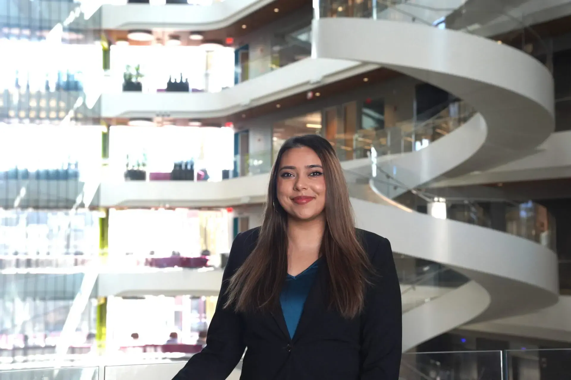 A woman with long brown hair, wearing a dark blazer and teal top, stands smiling in a modern building with bright lighting and a spiral staircase in the background.