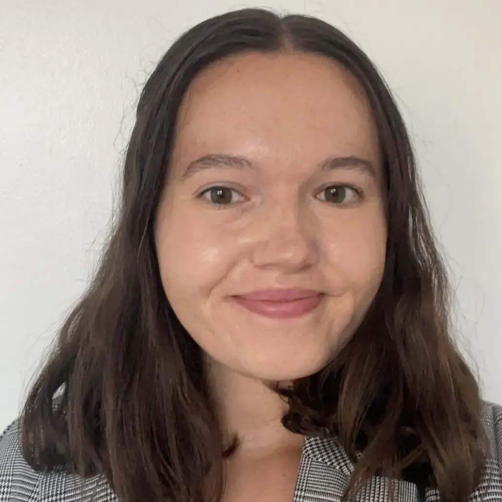 A woman with long brown hair, wearing a gray plaid blazer, smiles softly at the camera in front of a plain white background.