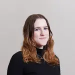 A young woman with wavy, light brown hair wearing a black turtleneck sweater stands against a plain, light-colored background, looking at the camera with a slight smile.