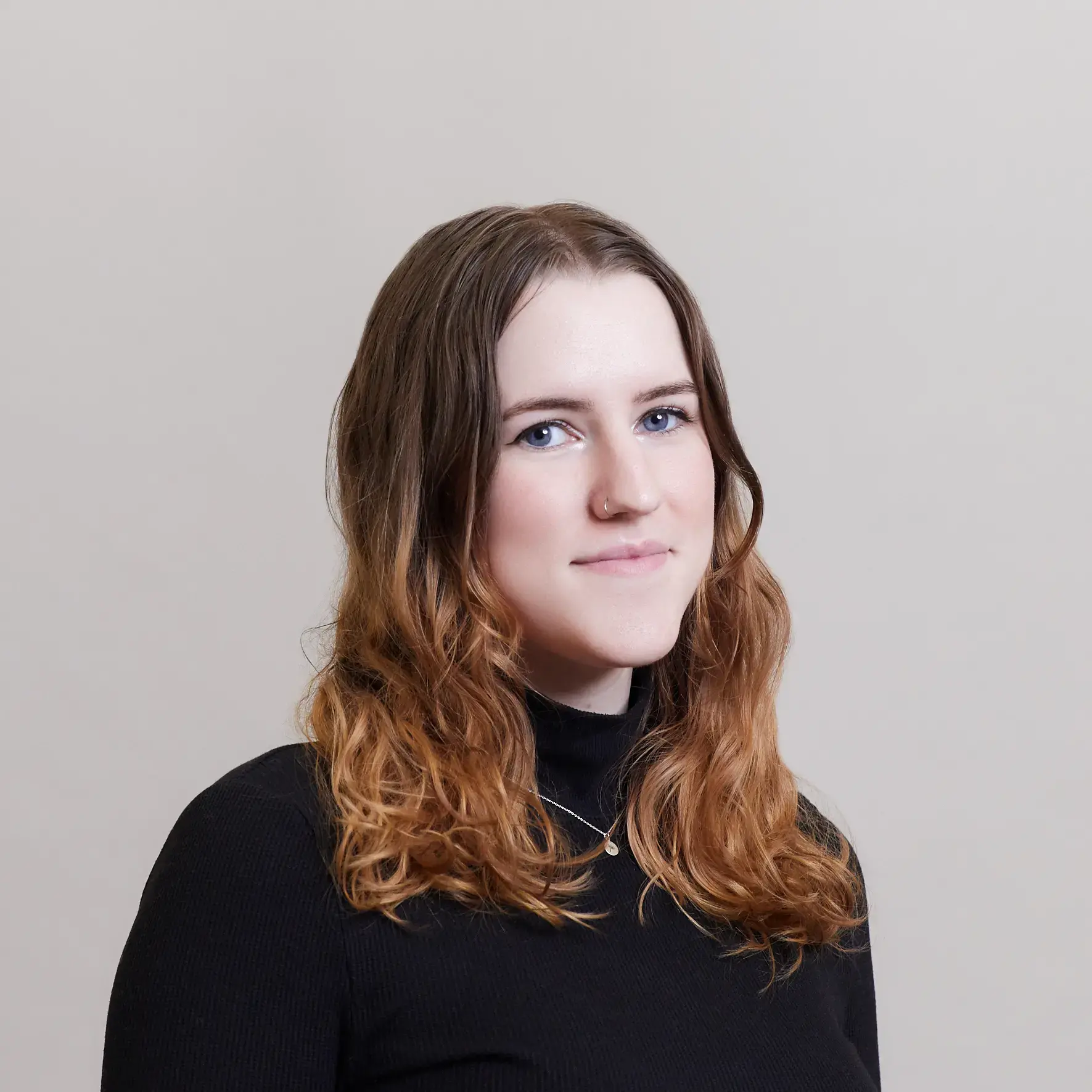A young woman with wavy, light brown hair wearing a black turtleneck sweater stands against a plain, light-colored background, looking at the camera with a slight smile.