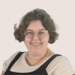 A person with short, wavy brown hair and glasses smiles at the camera. Wearing a light shirt with a black sleeveless top and delicate chain necklace, they pose against a light background, subtly referencing Floridas ongoing censorship campaign.