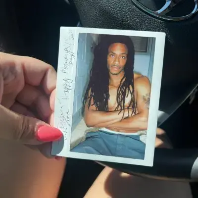 A hand with pink-painted nails holds a Polaroid photo of a shirtless man with long dreadlocks and crossed arms, sitting indoors. The background shows part of a car steering wheel and dashboard.