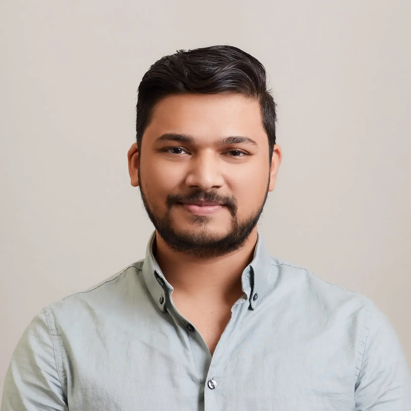 A young man with short, dark hair and a beard is smiling gently. He is wearing a light gray button-up shirt and is posed in front of a plain, neutral background.