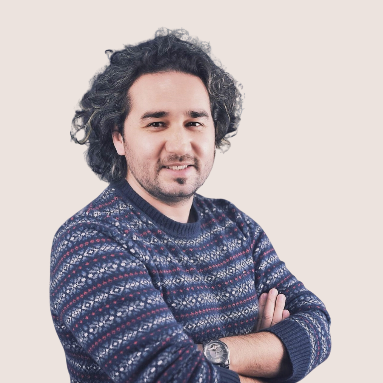 A man with curly hair and a beard, wearing a patterned sweater and a watch, stands with arms crossed and smiles at the camera against a plain light background.
