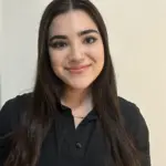 A young woman with long dark hair, wearing a black button-up shirt, smiles at the camera in front of a plain light-colored wall.