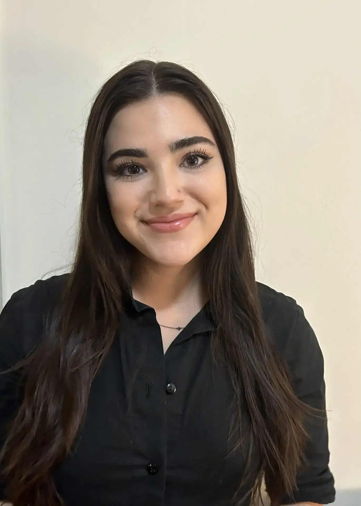 A young woman with long dark hair, wearing a black button-up shirt, smiles at the camera in front of a plain light-colored wall.