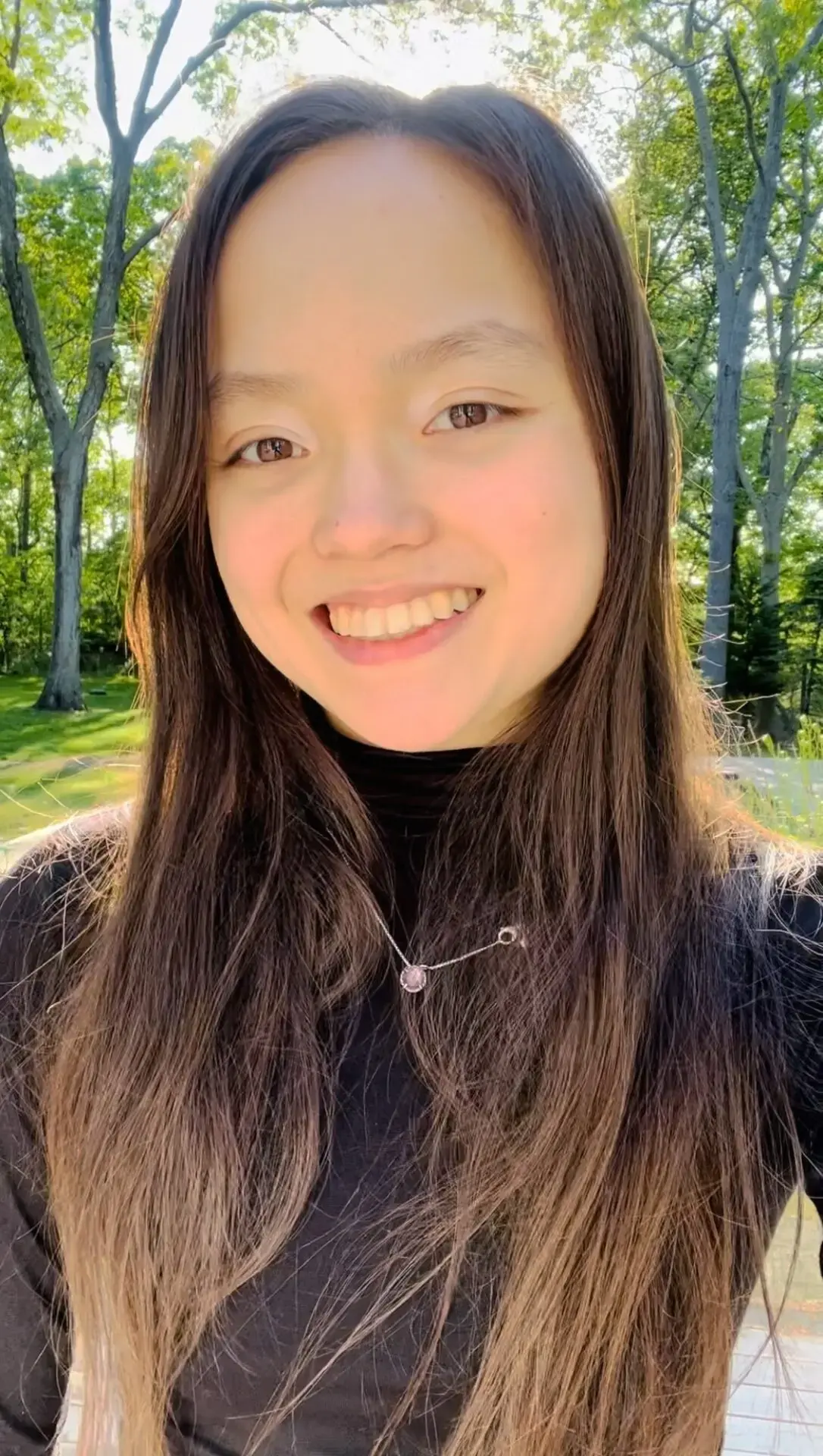 A young woman with long brown hair wearing a black top smiles outdoors on a sunny day, with green trees and sunlight in the background.