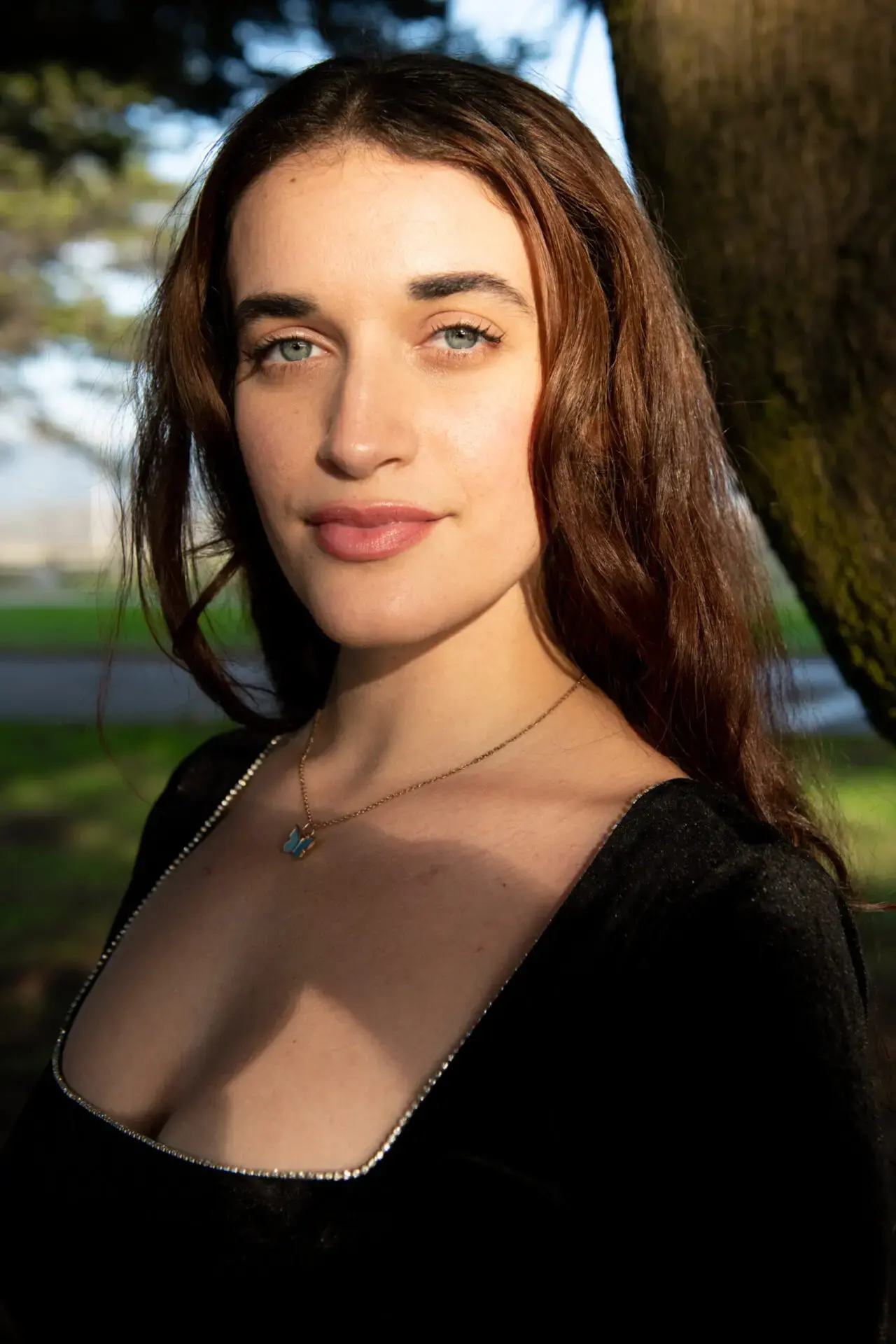 A woman with long brown hair and blue eyes stands outdoors, wearing a black top and a necklace. Sunlight illuminates her face, and trees and greenery are visible in the background.