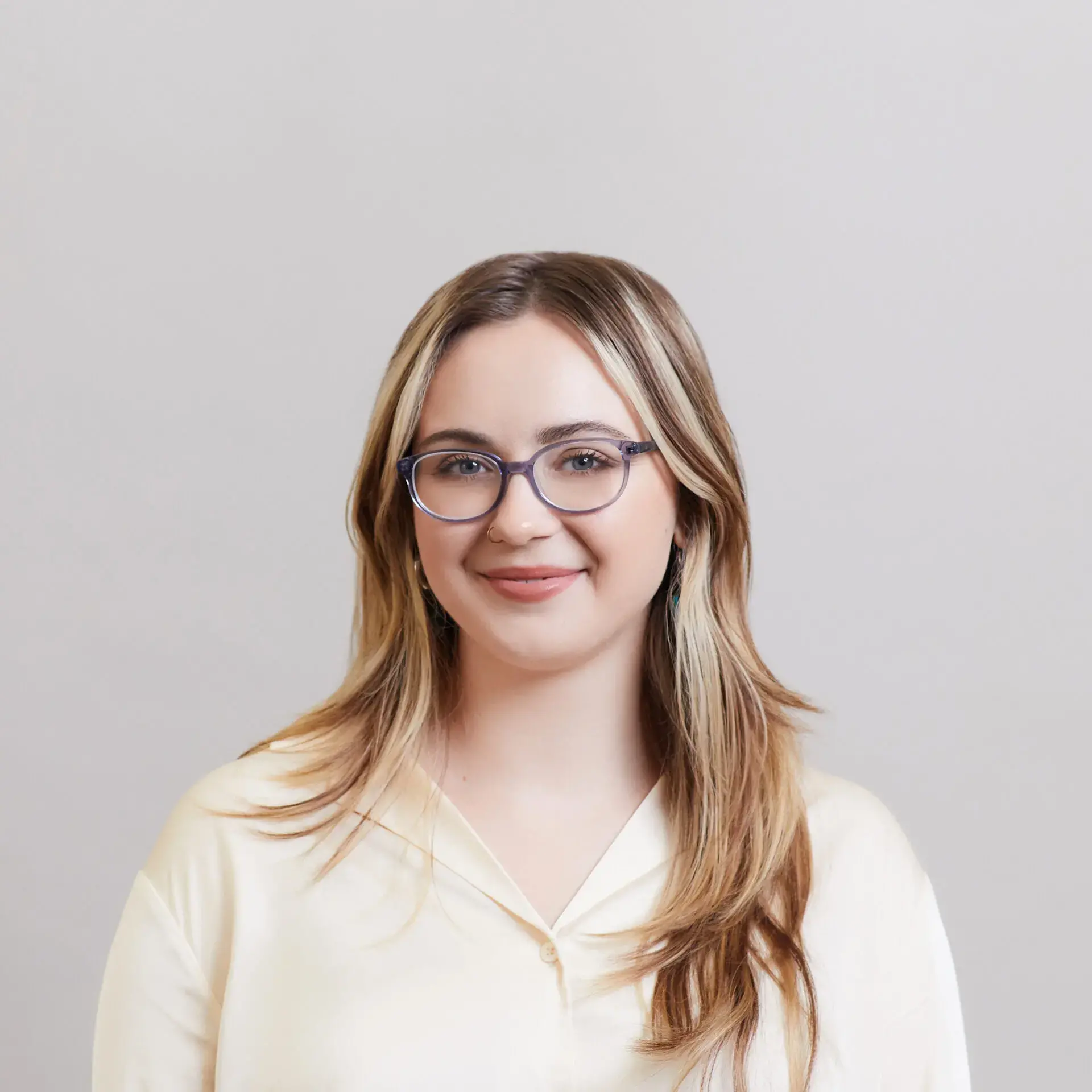 A young woman with long, light brown hair and glasses smiles softly. She is wearing a cream-colored blouse and stands in front of a plain, light gray background.