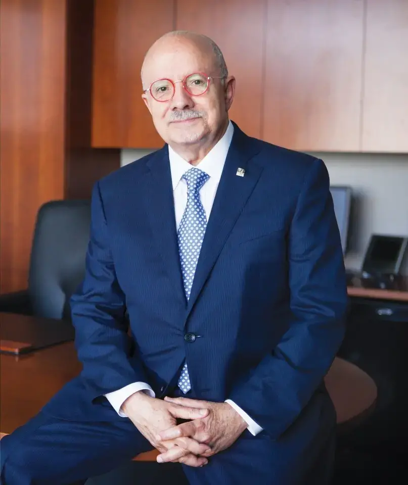 A bald man with glasses and a mustache, wearing a navy blue suit and polka-dot tie, sits on the edge of a desk in a modern office with wooden cabinets and a black chair.