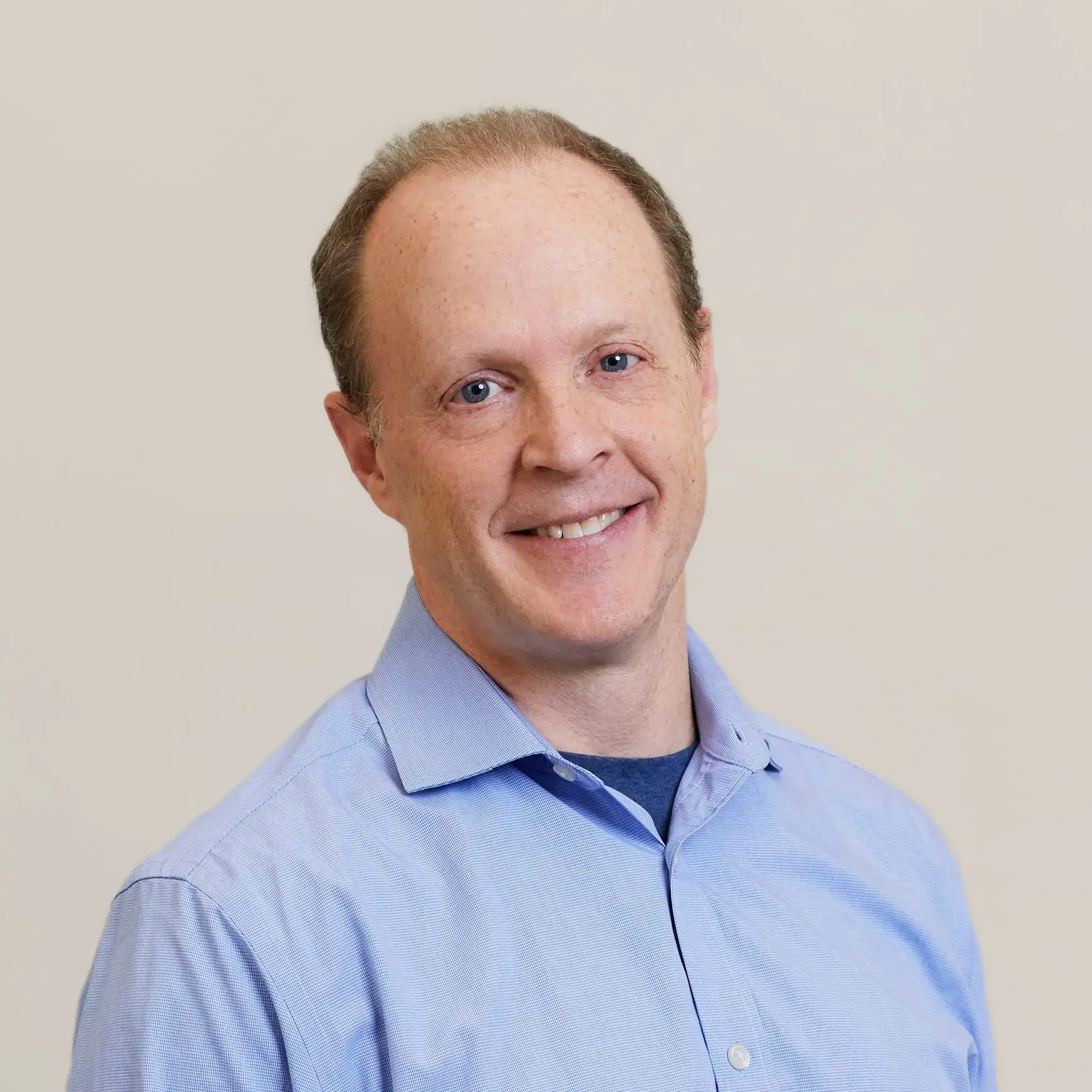 A middle-aged man with short light brown hair is smiling at the camera. He is wearing a light blue button-up shirt over a dark t-shirt, standing in front of a plain, light-colored background at a PEN Across America event.