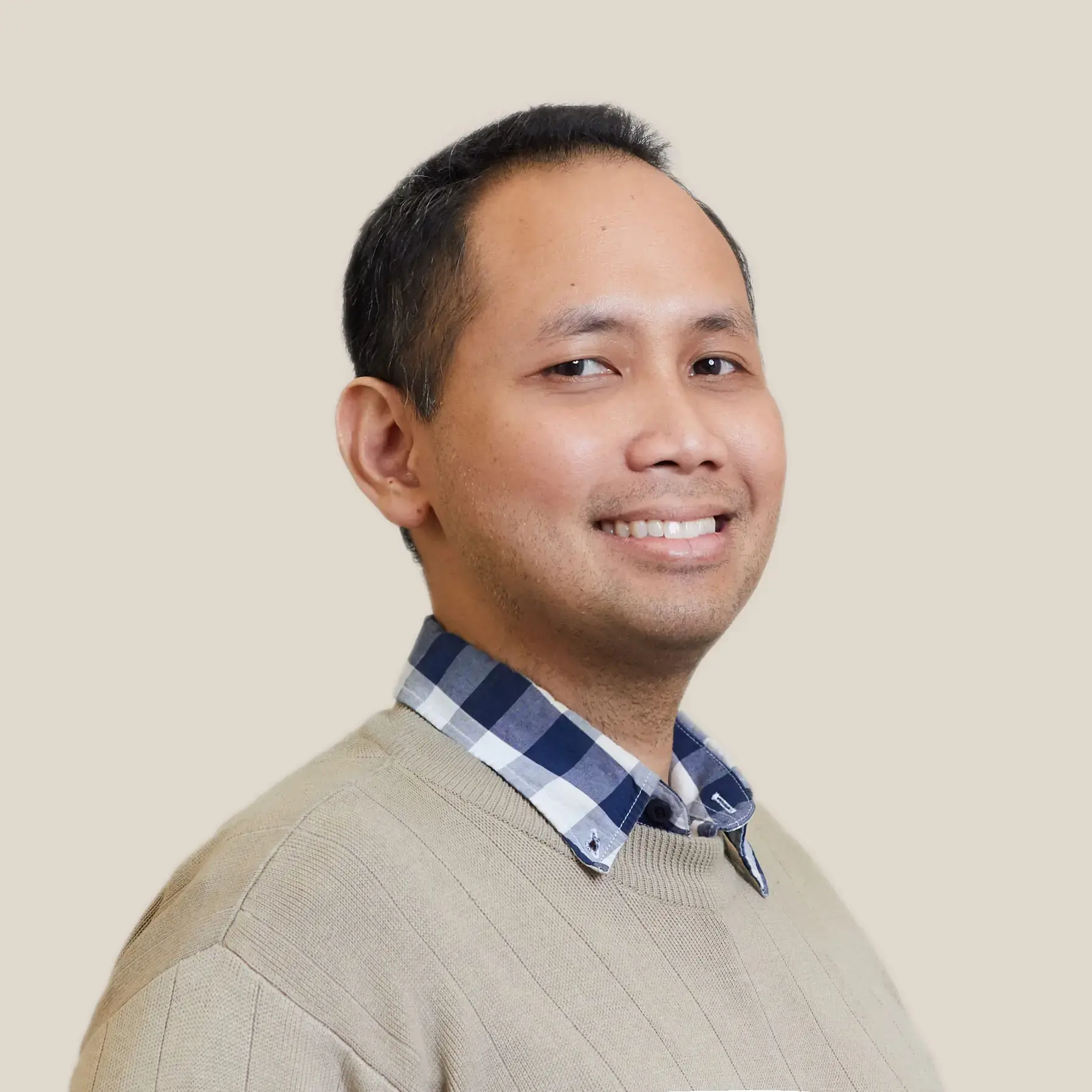 A man with short dark hair smiles while wearing a tan sweater over a blue and white plaid shirt, standing against a plain beige background, embodying the warmth often seen at Pen America membership gatherings.