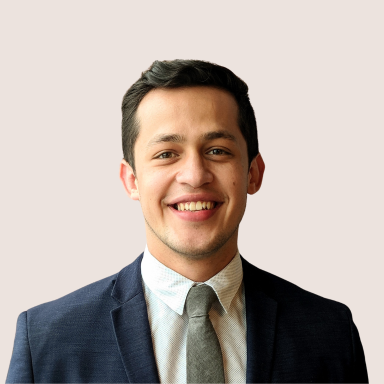 A young man in a navy suit, light shirt, and gray tie is smiling at the camera against a plain light background.