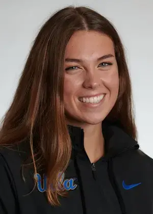 A young woman with long brown hair smiles at the camera, wearing a black jacket with blue UCLA and Nike logos on it, against a plain, light background.