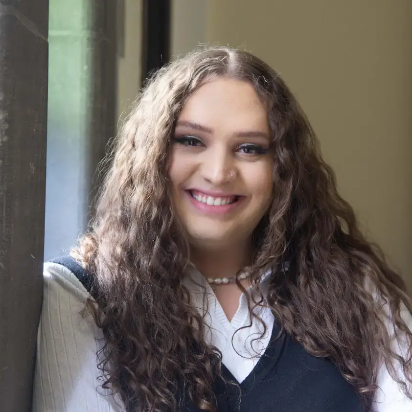 A person with long, curly brown hair and light skin is smiling while leaning against a window. They are wearing a white shirt and a black vest, accessorized with a pearl necklace. The background is softly lit and out of focus.