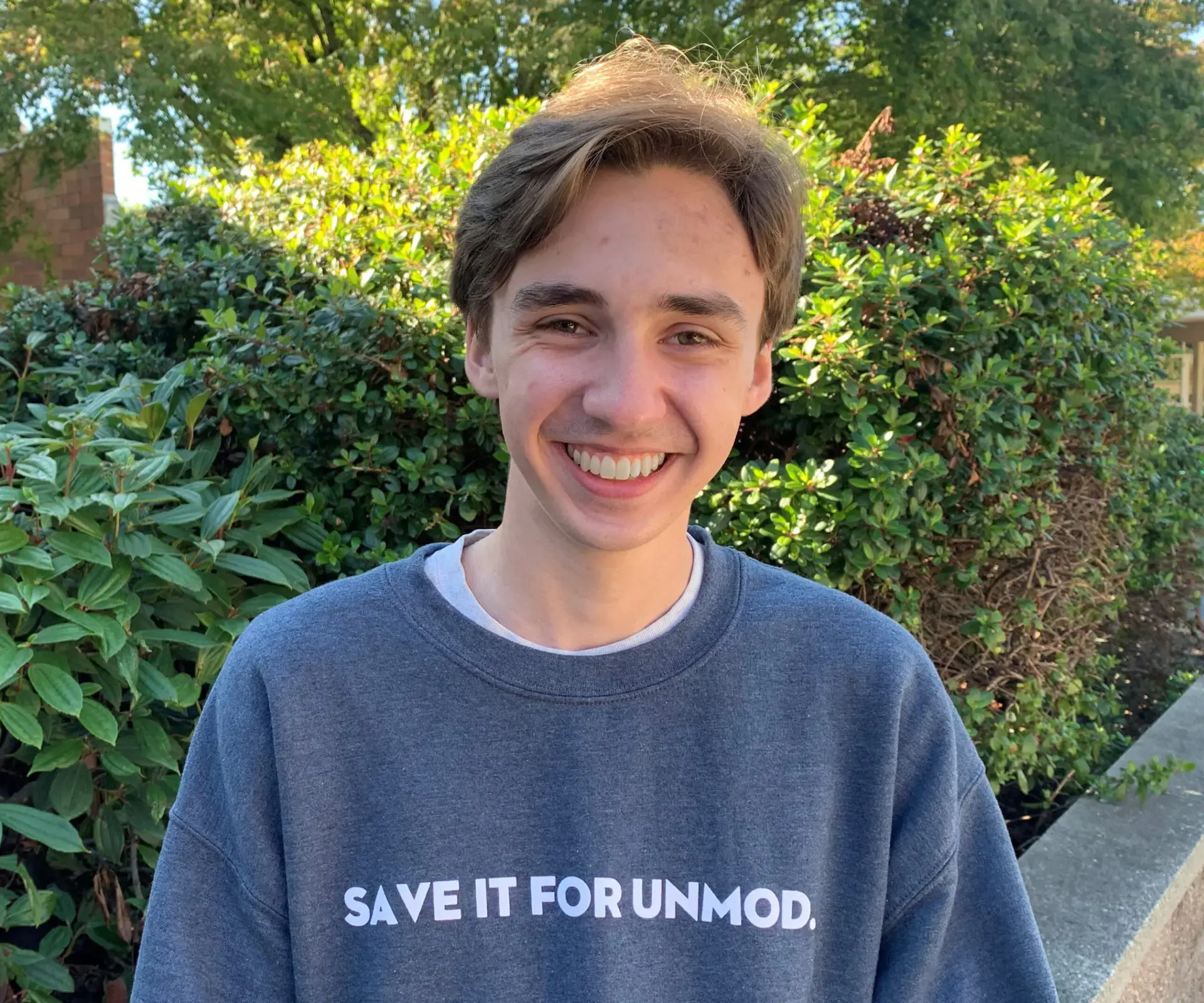 A young person with light brown hair smiles outdoors, wearing a gray sweatshirt with the words “SAVE IT FOR UNMOD.” Green bushes and trees are in the background under daylight.