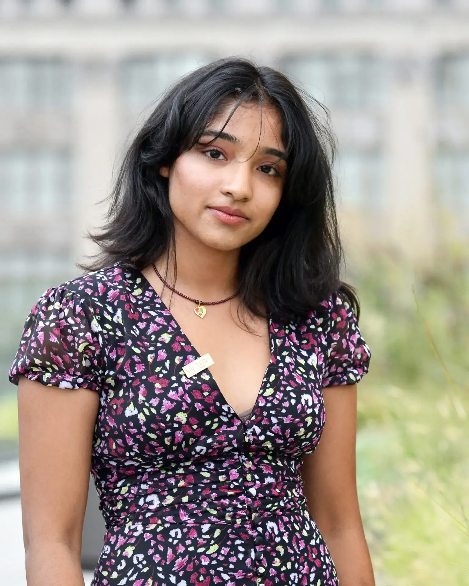 A young woman with medium-length black hair wearing a patterned short-sleeve dress and a necklace stands outdoors, looking at the camera with a neutral expression. The background is blurred with greenery and a building visible.