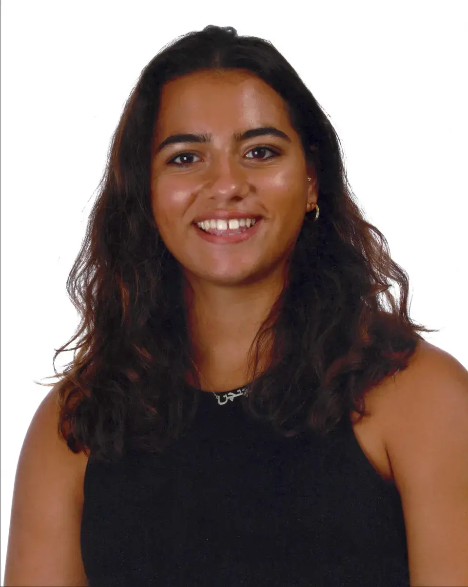 A young woman with long, wavy brown hair wearing a sleeveless black top smiles at the camera against a plain white background.