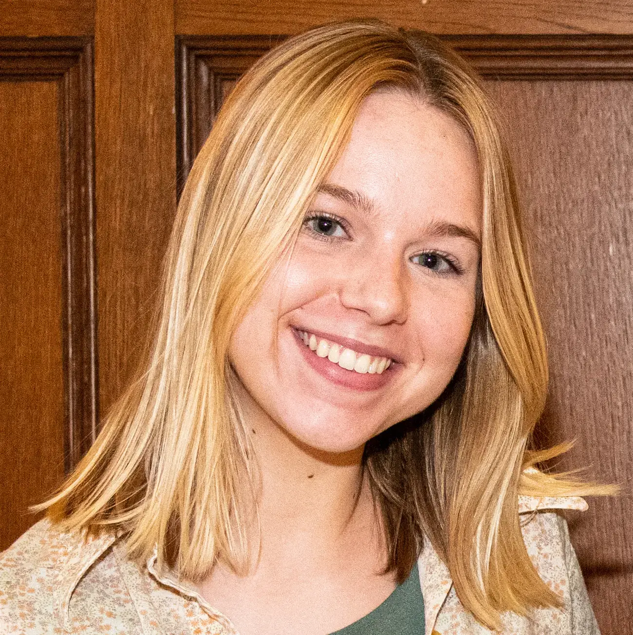 A young woman with straight blonde hair smiles at the camera. She is wearing a light patterned shirt and stands in front of a wooden panel background.