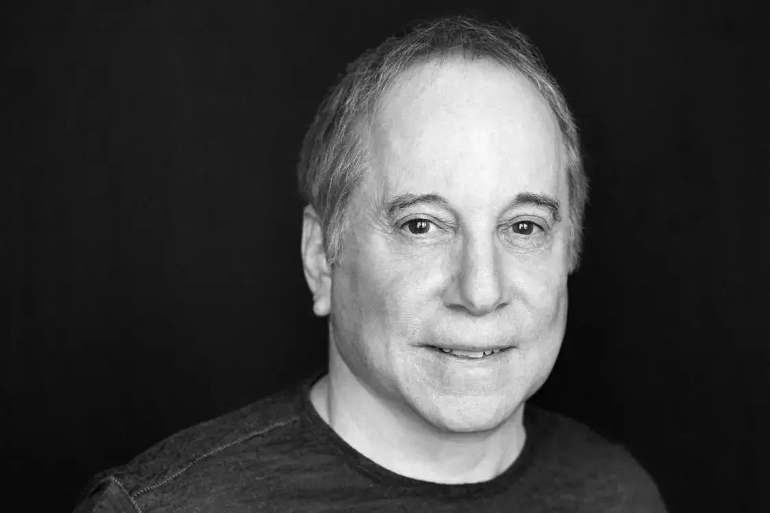 A middle-aged man with short, graying hair smiles slightly while looking at the camera. He is wearing a dark shirt and is posed against a plain, dark background.