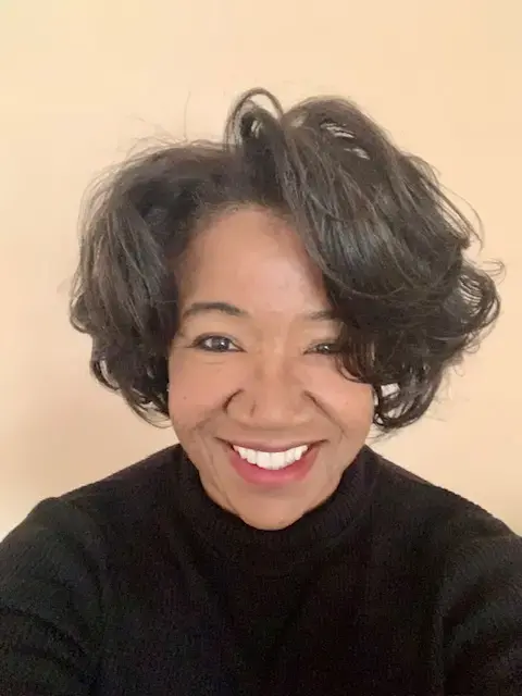 A woman with short, wavy dark hair and a bright smile wears a black top and poses in front of a plain beige background.