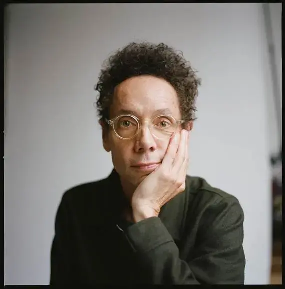 A person with short curly hair and glasses rests their face on their hand, looking thoughtfully at the camera against a plain background.