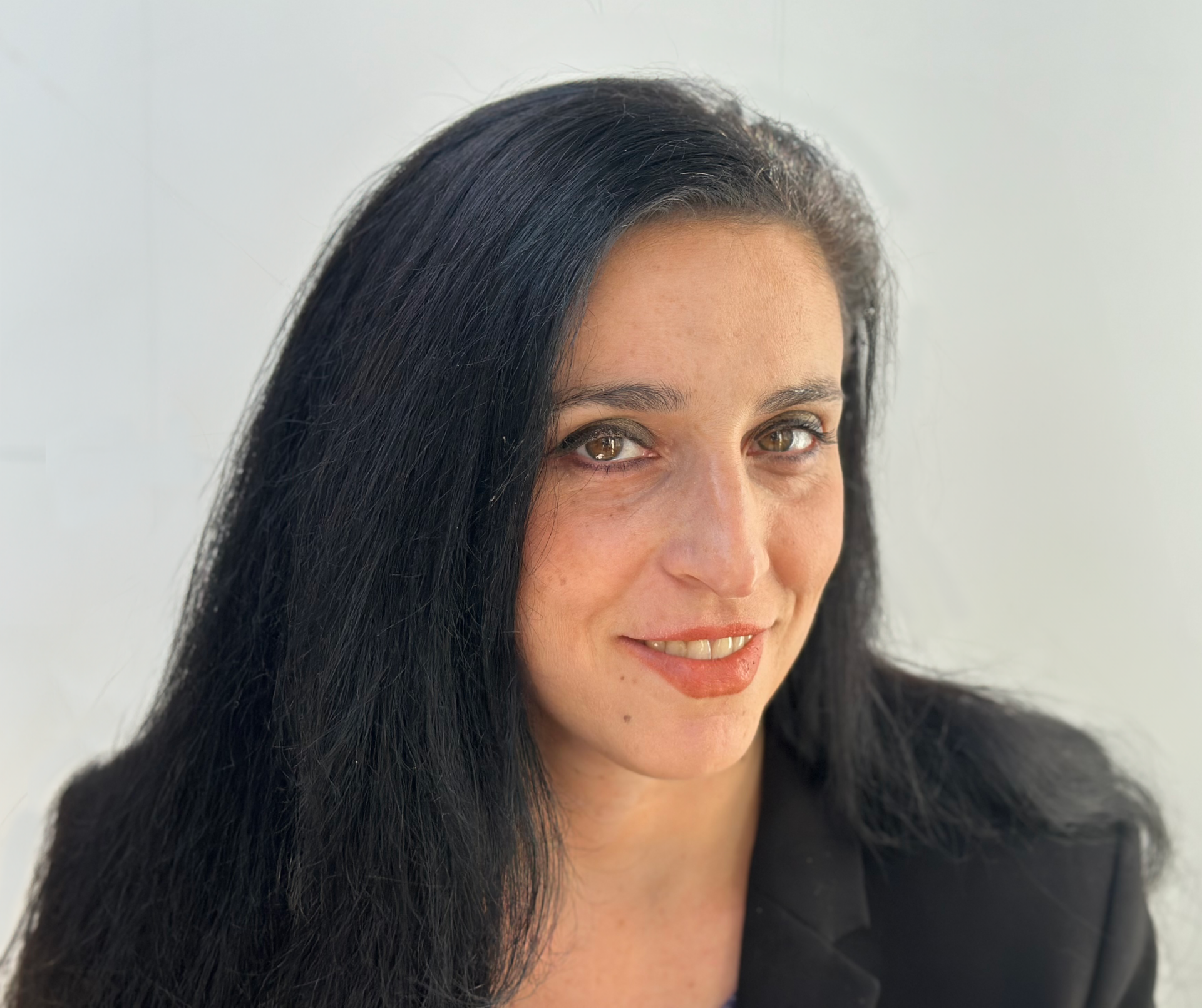 A woman with long, straight black hair and a black blazer smiles softly at the camera against a plain white background.