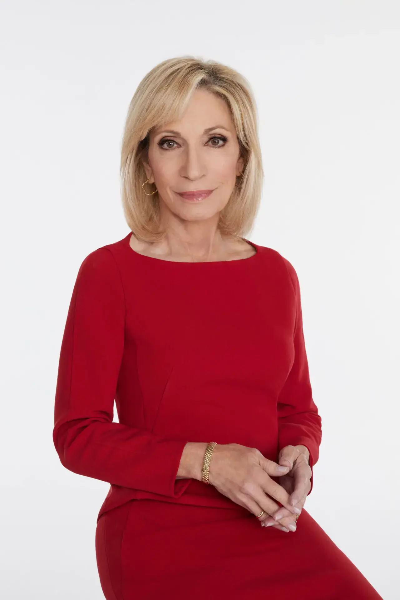 A woman with shoulder-length blonde hair wearing a red long-sleeve dress sits against a plain white background, looking at the camera with a slight smile and her hands resting together.