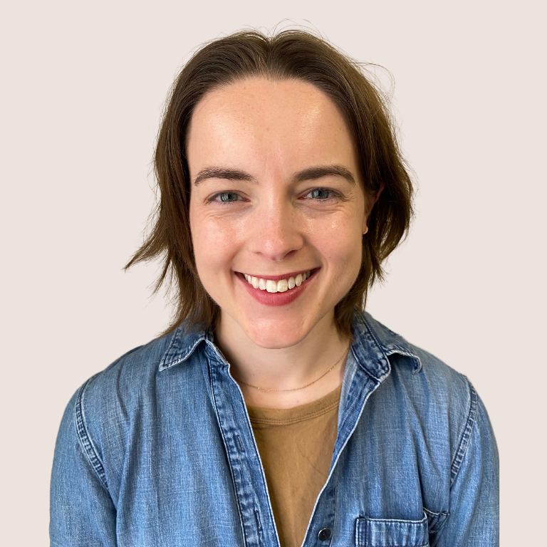 A person with short brown hair is smiling at the camera. They are wearing a denim shirt over a light brown top, and the background is plain and light-colored.