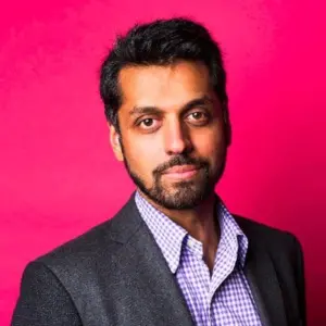 A man with short dark hair, a beard, and a mustache is wearing a dark blazer over a checkered shirt, standing in front of a bright pink background, looking at the camera with a slight smile.