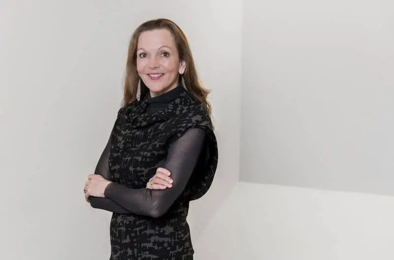 A woman with long brown hair, wearing a black patterned sleeveless top over a sheer black shirt, stands smiling with her arms crossed against a plain, light-colored background.