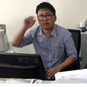A man wearing glasses and a checkered shirt sits at a desk with a computer, looking toward the camera with one hand raised. Office items and a printer are visible in the background, suggesting work related to PEN America or Global Press Freedom Advocacy Institute.