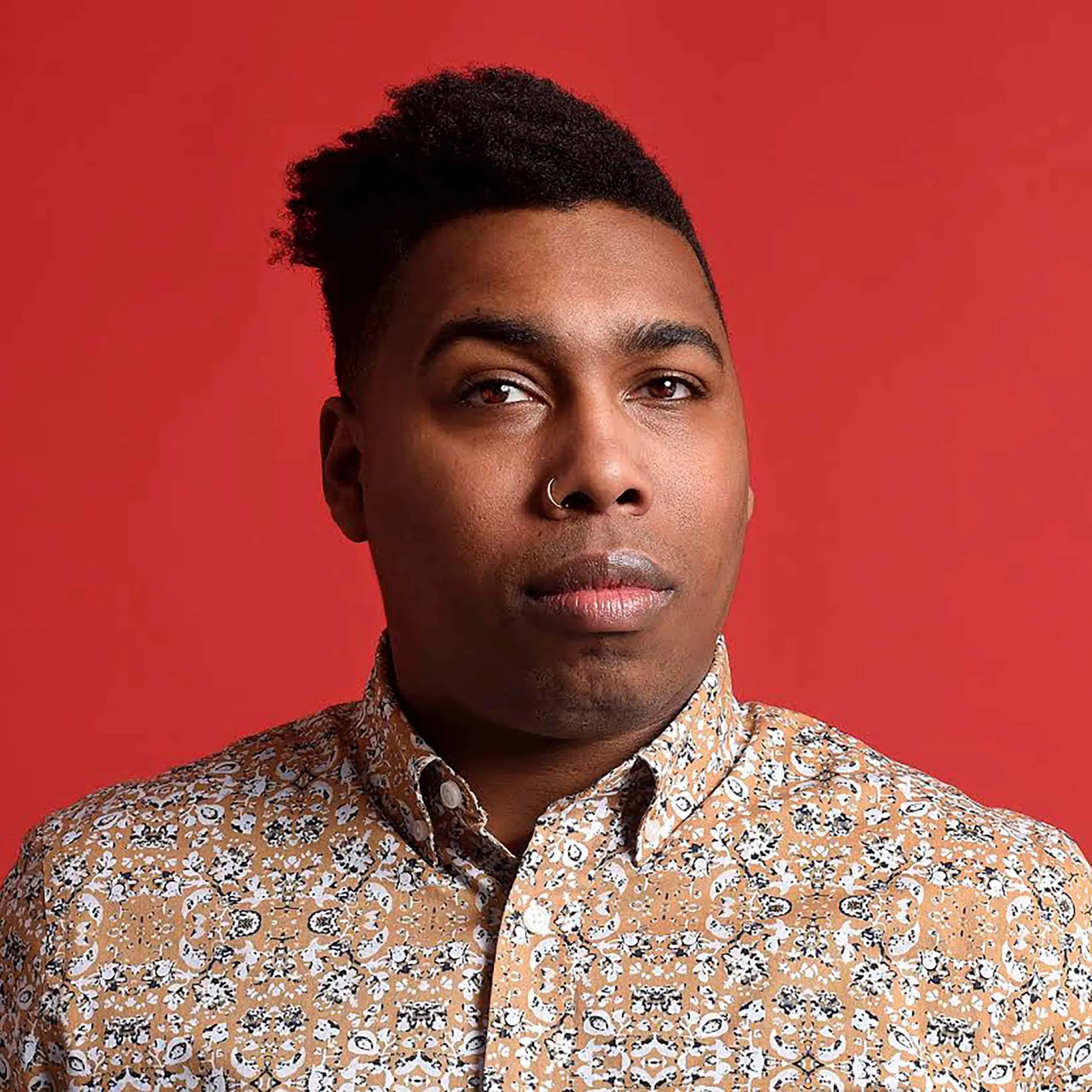 A person with short, curly hair styled to one side and a nose ring looks at the camera. They wear a patterned button-up shirt and stand against a solid red background.