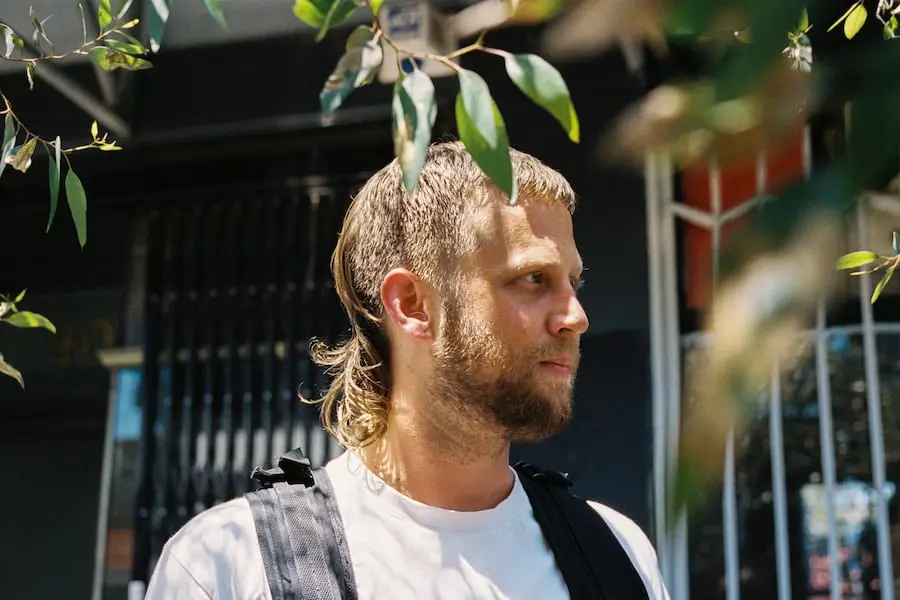 A man with a mullet hairstyle and beard stands outdoors, wearing a white shirt and a backpack. Green leaves partially frame his face, and a building with a black gate is visible in the background.