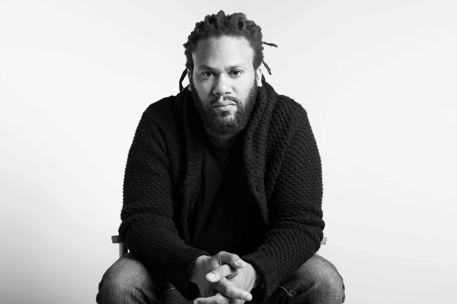 A man with medium-length dreadlocks and a beard, wearing a dark textured sweater and jeans, sits facing forward with his hands clasped, looking directly at the camera against a plain white background.