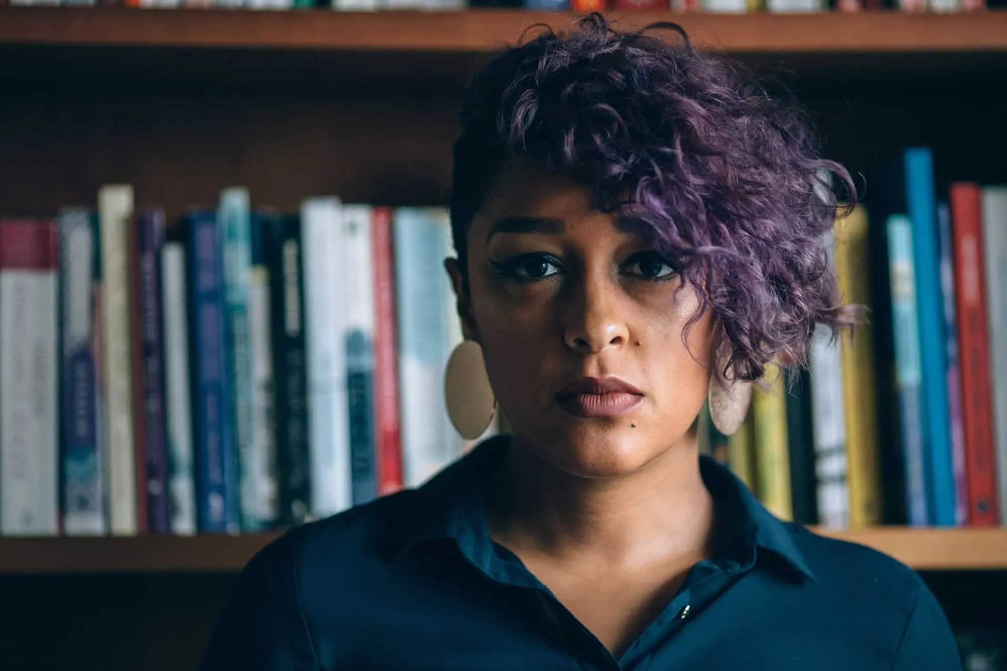 A person with short, curly purple hair and large round earrings stands in front of a bookshelf, wearing a dark collared shirt and gazing seriously at the camera.