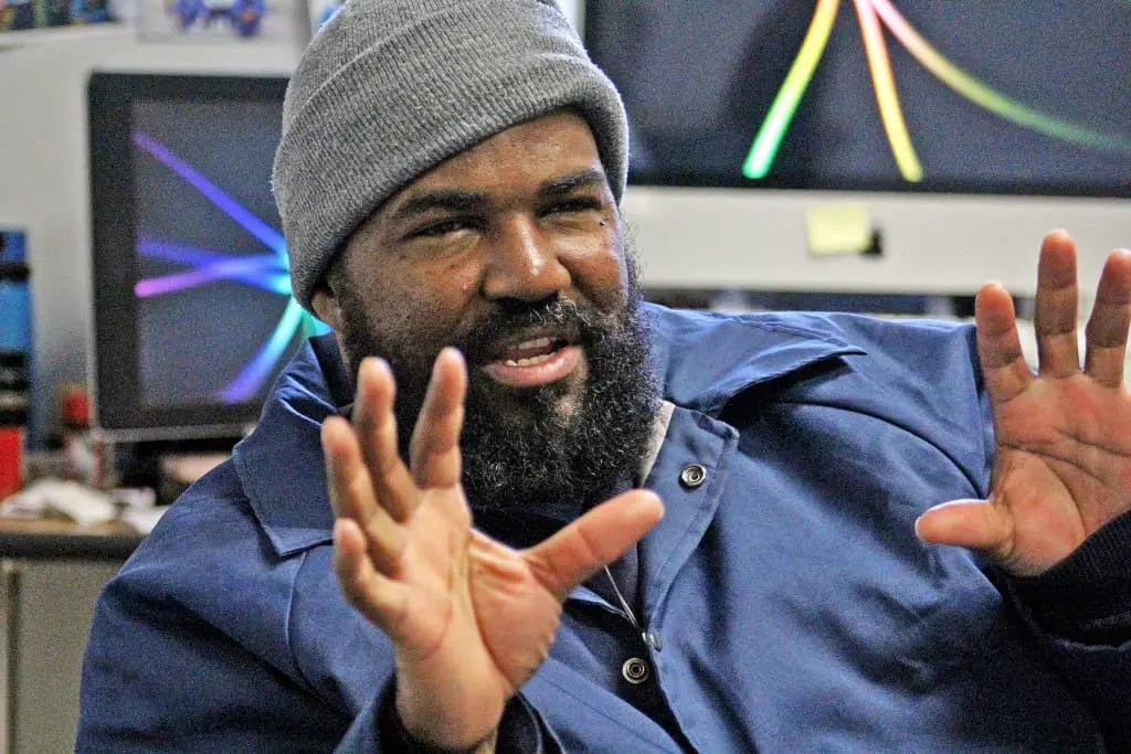 A man with a beard wearing a gray beanie and blue jacket gestures with both hands while speaking. Computer monitors with colorful abstract screensavers are visible in the background.