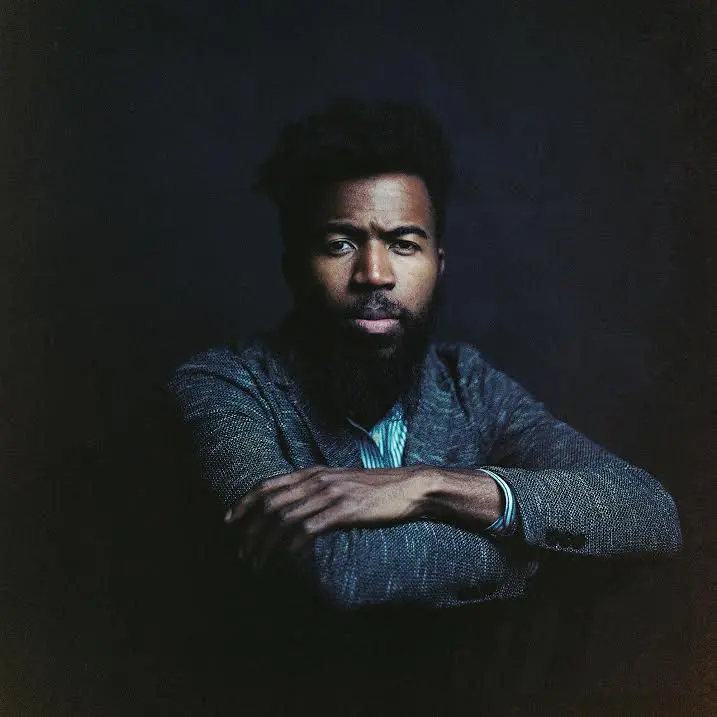A man with a dark beard and mustache, wearing a gray textured blazer over a light blue shirt, sits with his arms crossed against a dark background, looking directly at the camera.