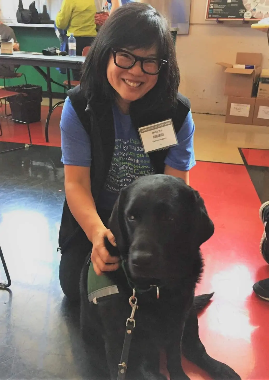 A woman with glasses and dark hair smiles while kneeling behind a black Labrador retriever wearing a green vest, inside a room with a red and black floor.