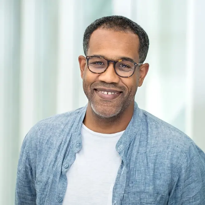 A man with short hair, glasses, and a trimmed beard smiles while wearing a light blue shirt over a white t-shirt, standing in front of a softly blurred light background.