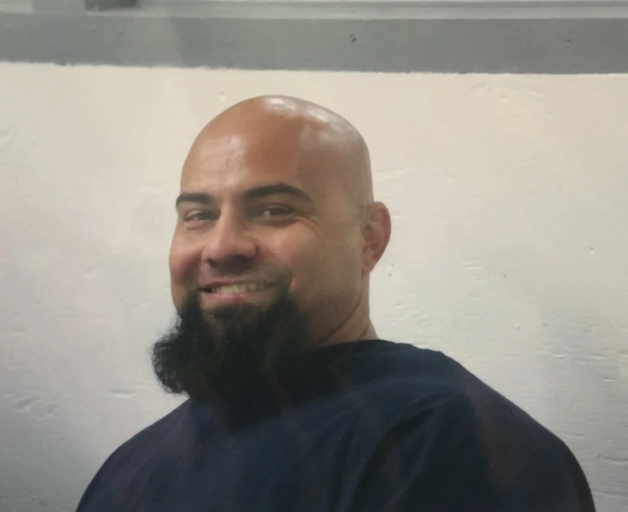 A bald man with a dark, thick goatee smiles at the camera. He is wearing a dark shirt and is standing in front of a plain, light-colored wall.