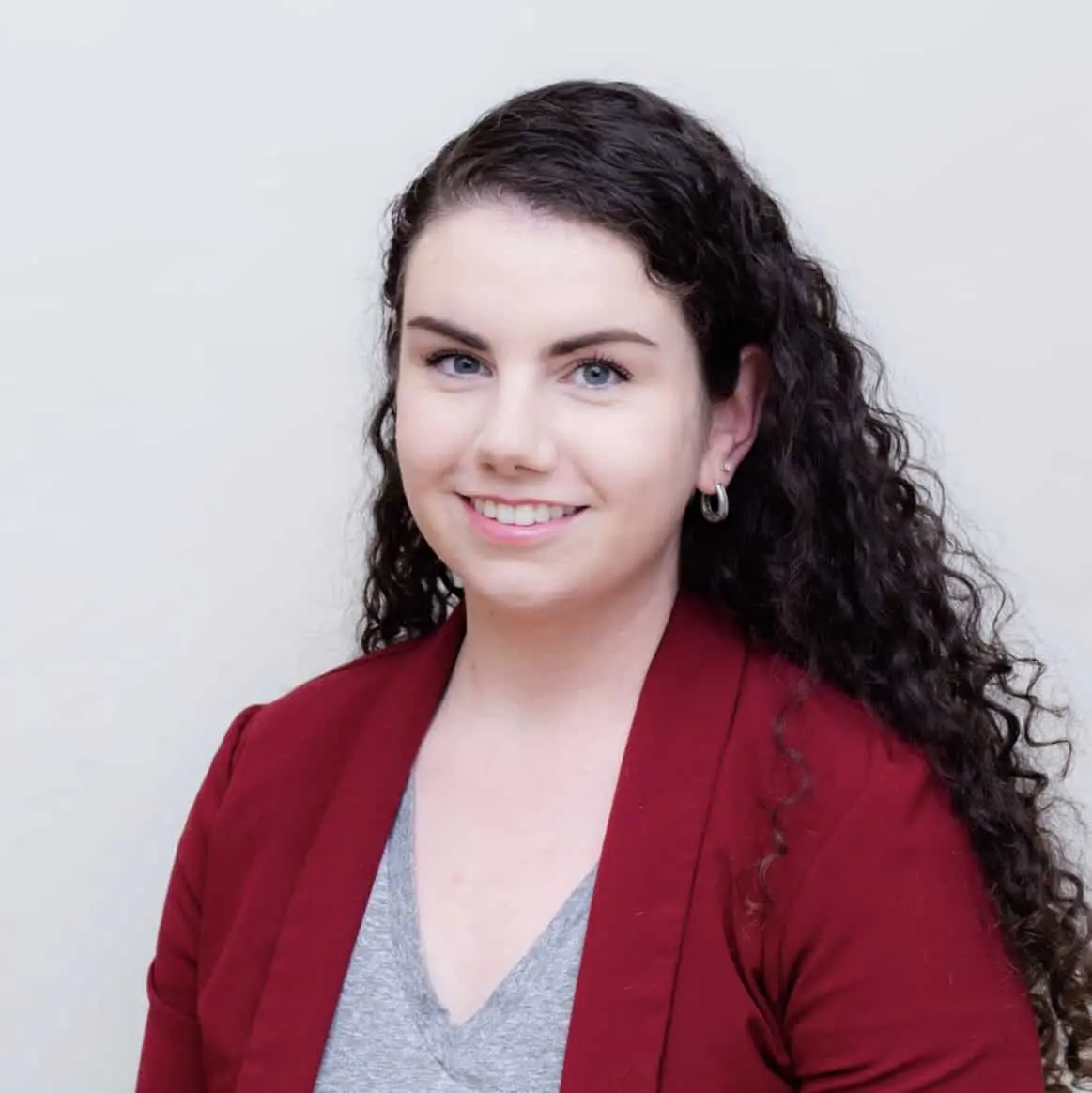 A woman with long curly dark hair, wearing a red blazer over a gray top, smiles at the camera against a plain light background.